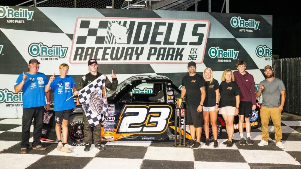 Racing team celebrating a victory lane win at Dells Raceway Park, gathered around truck #23 with a checkered flag and trophy, sponsored by O'Reilly Auto Parts.