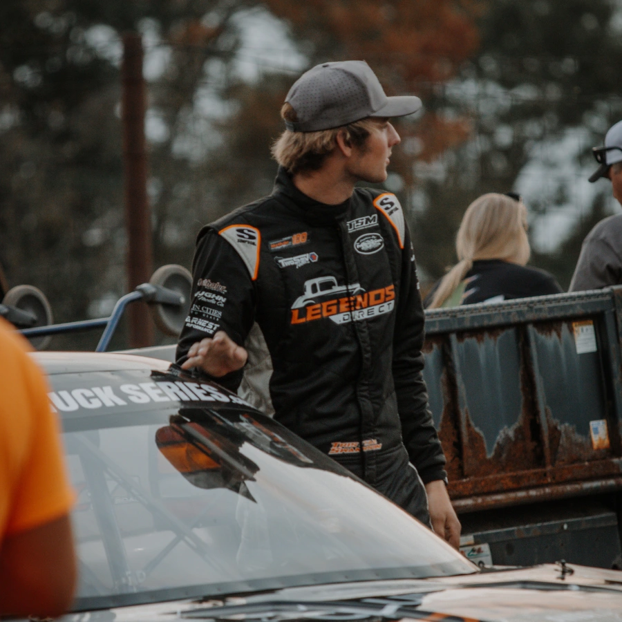 Race driver in Legends Direct fire suit standing beside a Truck Series race car in the paddock