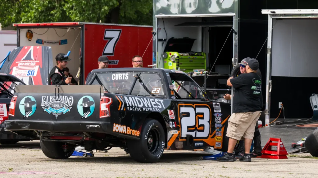 Race truck #23 sponsored by Midwest Racing Network and El Bandido Yankee parked in the pit area at a short-track racing event, with crew members and enclosed haulers visible in the background.