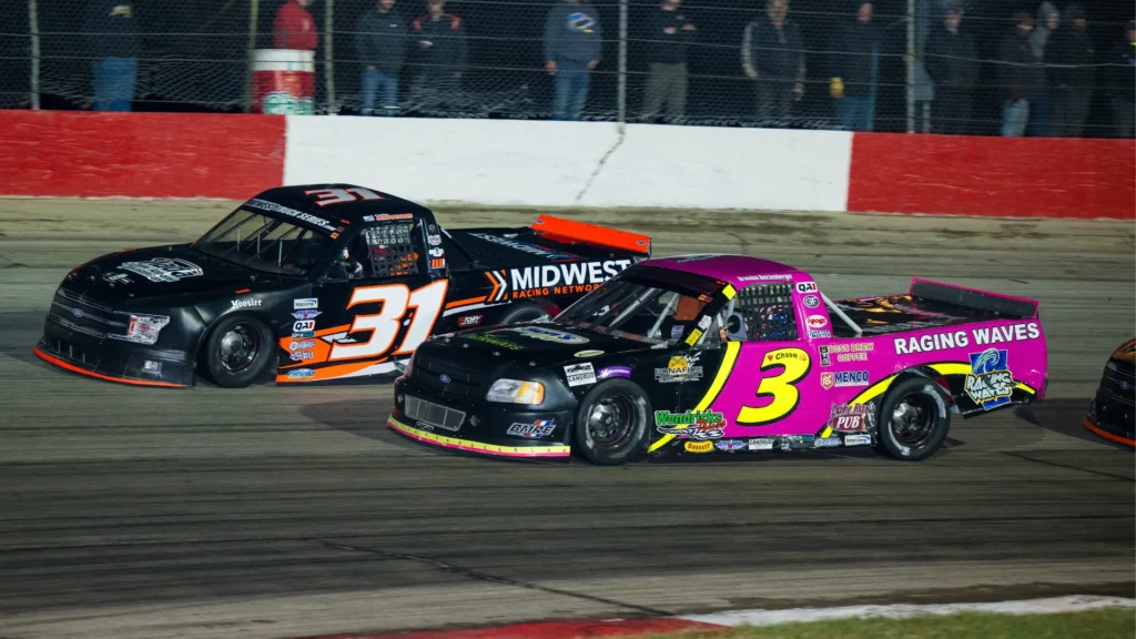 Midwest Truck Series truck #31 with Midwest Racing Network sponsorship racing side by side against truck #3 with Raging Waves sponsorship on an illuminated short-track oval at night with fans visible in the grandstands.